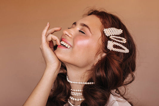 Woman wearing pearl jewelry and hair clips, smiling joyfully against a brown background.